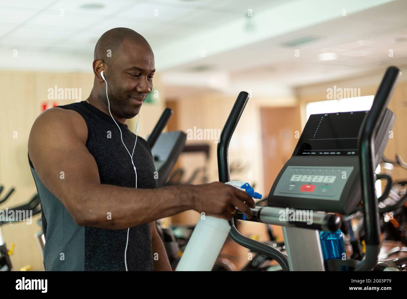 Schwarzer männlicher Athlet, der im Fitnessstudio ein Herz-Kreislauf-Gerät mit Desinfektionsmittel reinigt. Selektiver Fokus. Stockfoto