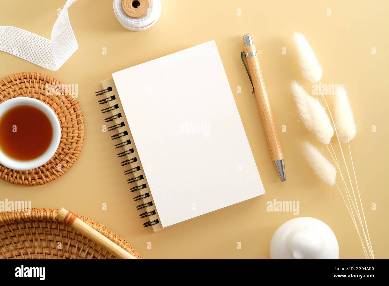 Bastelpapier-Notizblock mit leerem Papiernotiz, Bleistift, Rattanplatte, Kaffeetasse, Band, Getrocknete Blüten auf beigem Hintergrund. Flach liegend, Draufsicht. Elegantes fe Stockfoto