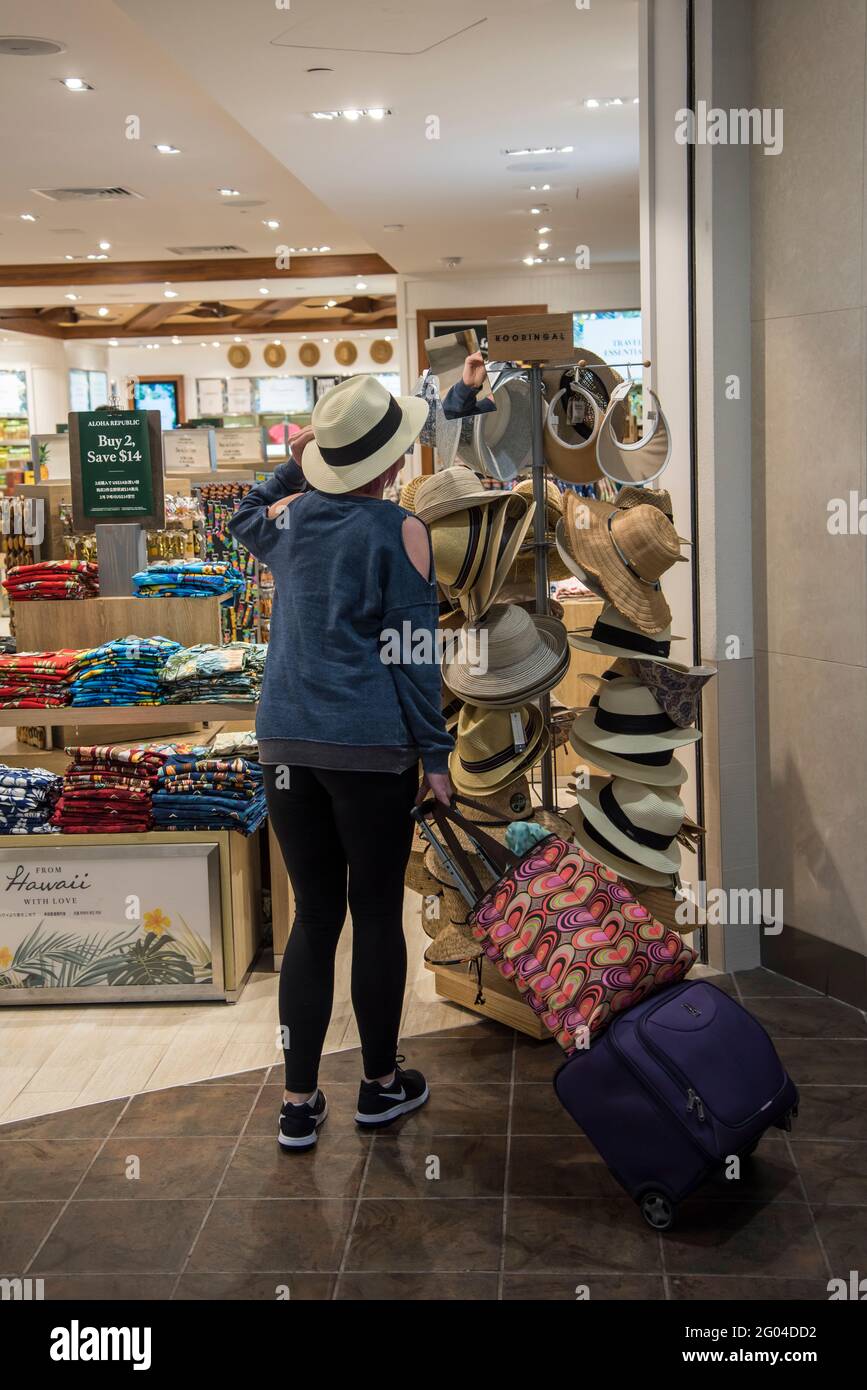 Honolulu, Hawaii. Eine Frau mittleren Alters, die in einem Geschäft am Flughafen Hüte anprobiert, während sie während eines Besuchs auf Hawaii auf ihr Flugzeug wartet. Stockfoto