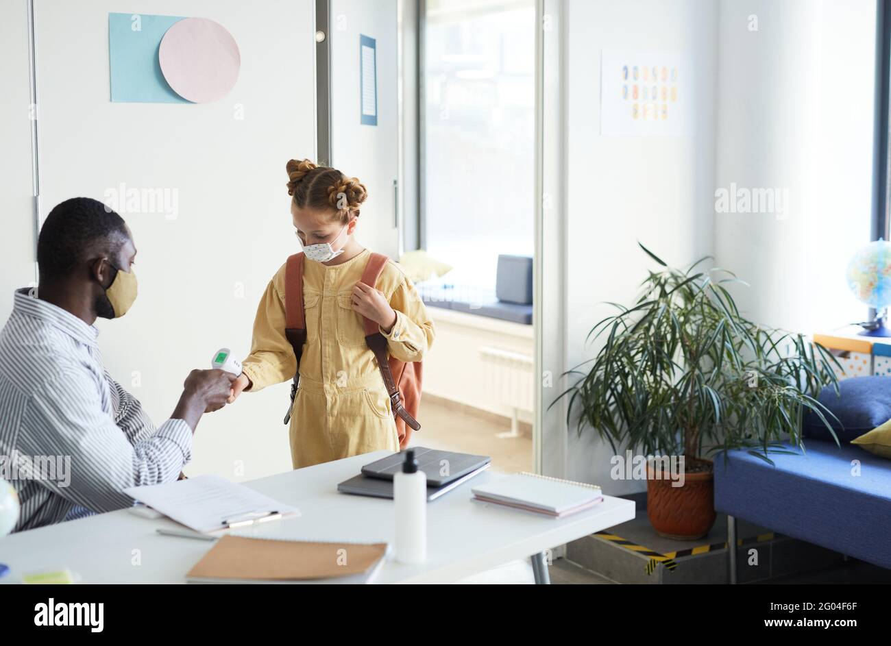 Porträt eines männlichen Lehrers, der die Temperatur des Mädchens überprüft, das in die Schule eingeht, und Sicherheitsmassnahmen Stockfoto