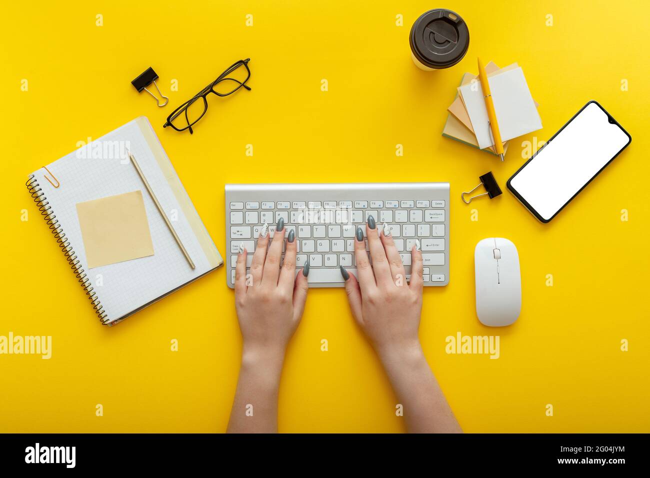 Frau, die am Arbeitsplatz auf der Computertastatur auf gelbem Hintergrund tippt. Büro-Desktop-Arbeitsplatz mit weiblichen Händen, Tasse Kaffee Smartphone Mockup Stockfoto