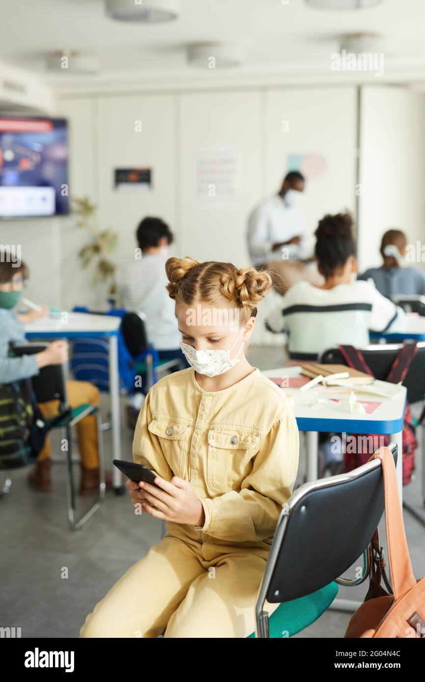 Vertikales Porträt von niedlichen Mädchen trägt Maske in der Schule und mit Smartphone, covid Sicherheitsmaßnahmen, kopieren Raum Stockfoto