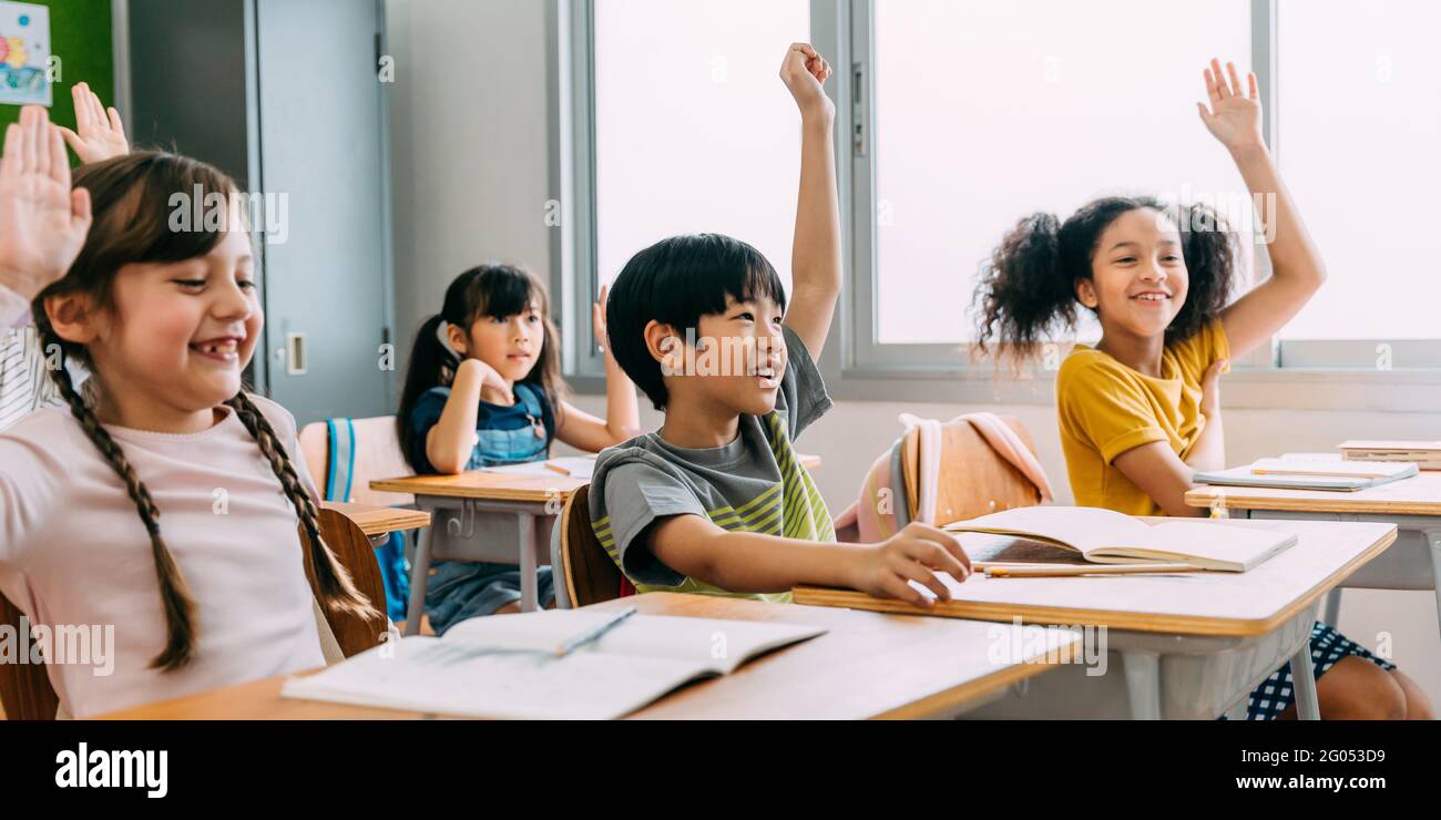 Im Grundschulalter hob der asiatische Student Junge in der Klasse die Hände hoch. Vielfältige Gruppe von Vorschulkindern im Grundschulalter in der Bildungsbauschule. Freiwilligenarbeit und Teilnahme im Klassenraum. Stockfoto