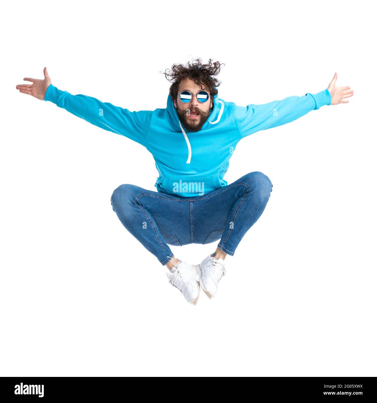 Ungeschoren Casual guy im Hoodie mit Sonnenbrille, die in der Luft springt und die Arme öffnet, während sie Spaß hat und vor weißem Hintergrund im Gestüt positiv ist Stockfoto