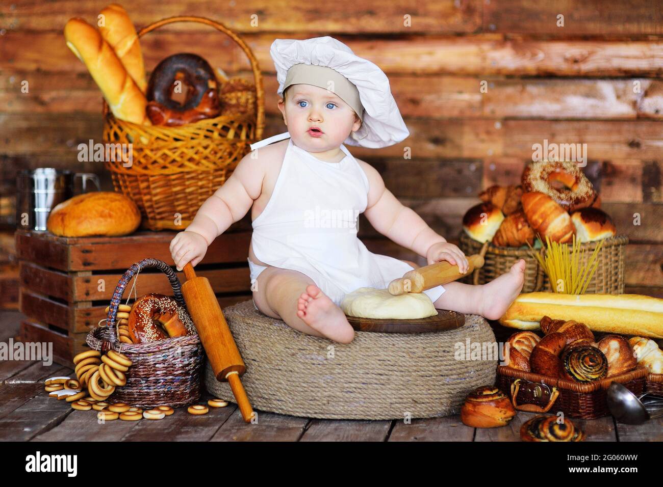 Der kleine Bäckerjunge mit Kochmütze und Schürze rollt rohen Teig mit einem hölzernen Nudelholz vor dem Hintergrund von Backwaren aus. Stockfoto