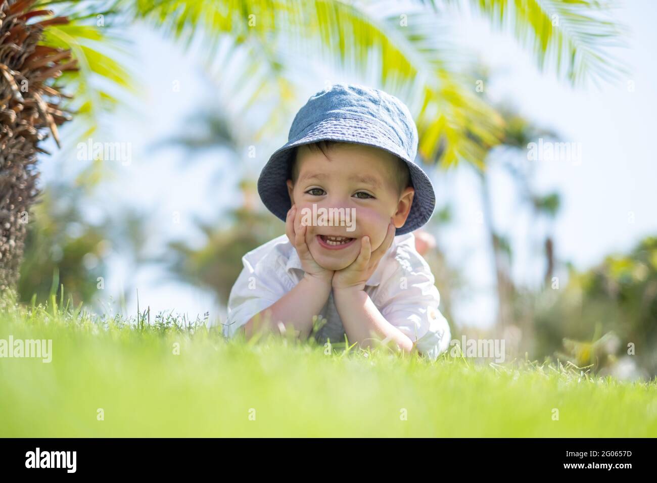 Kleiner Junge, der auf grünem Gras auf Rasen unter Palmen liegt. Blauer Himmel auf dem Hintergrund. Urlaub und glückliche Kindheit Konzept Stockfoto