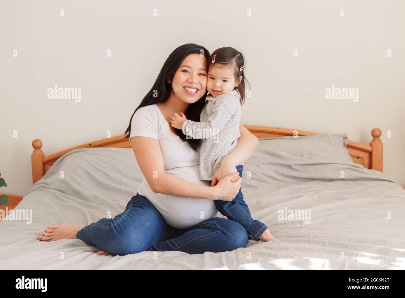 Asiatische chinesische schwanger Frau mit Kleinkind Mädchen umarmt auf dem Bett zu Hause. Mädchen Tochter Kind spielend mit Mutter. Mutter und kleine Tochter warten darauf, zu warten Stockfoto