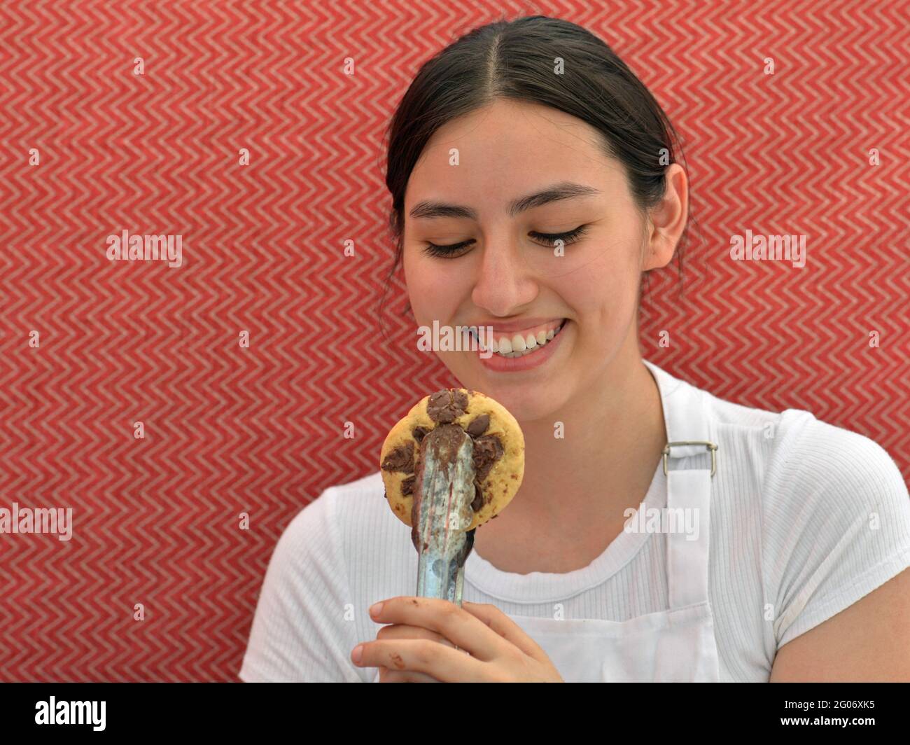 Junge charmante Brünette Verkäuferin lächelt und schaut auf einen frisch gebackenen Gourmet-Chocolate-Chip-Cookie in einer Essenzange vor einem roten Hintergrund. Stockfoto