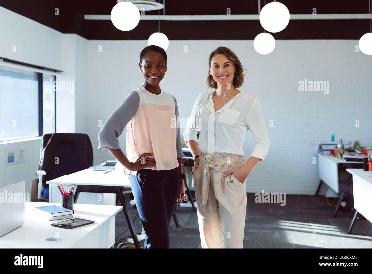 Zwei lächelnde, vielfältige Geschäftsfrauen stehen und blicken bei der Arbeit auf die Kamera Stockfoto
