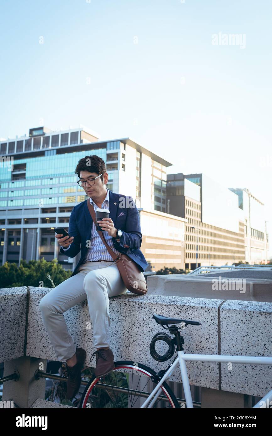 Asiatischer Geschäftsmann mit Smartphone hält Kaffee zum Mitnehmen an seinem Fahrrad in der Stadtstraße Stockfoto