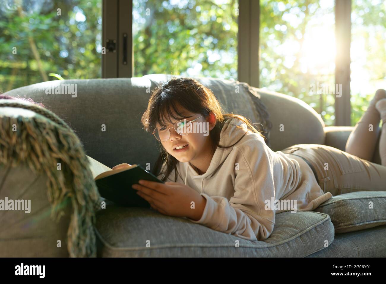 Lächelndes asiatisches Mädchen in einer Brille, das ein Buch liest und auf dem Sofa liegt Stockfoto