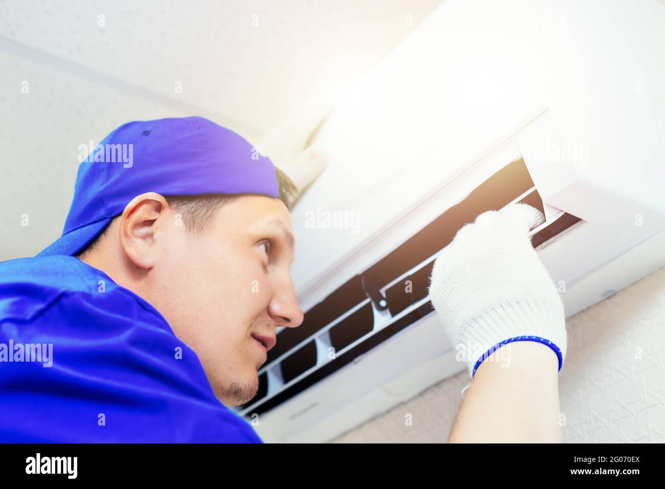 Ein junger Arbeiter mit blauer Baseballmütze und Handschuhen inspiziert und wartet die Klimaanlage im Zimmer. Porträt eines Technikers bei der Arbeit. Nahaufnahme. Modell Stockfoto
