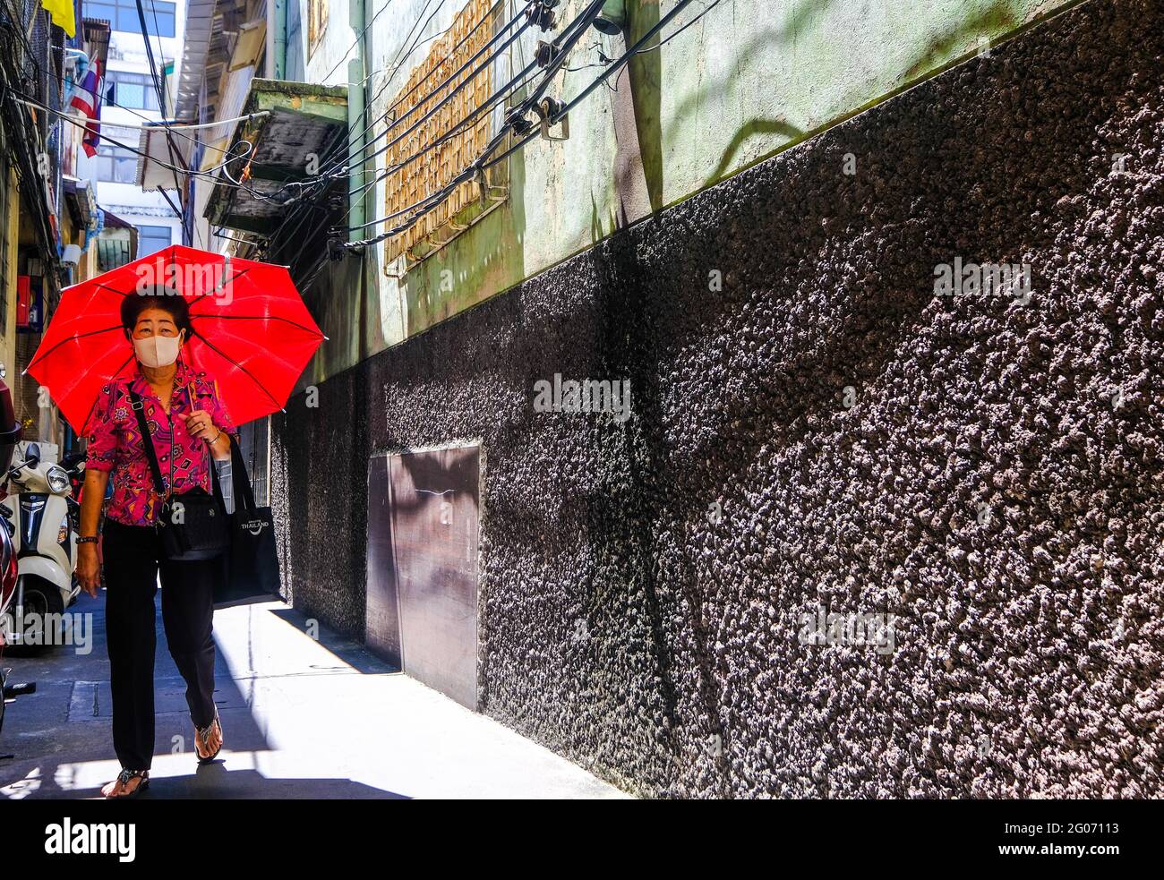 Eine Frau mit einem roten Regenschirm geht entlang einer sonnendurchfluteten Gasse in Chinatown, Bangkok, Thailand Stockfoto