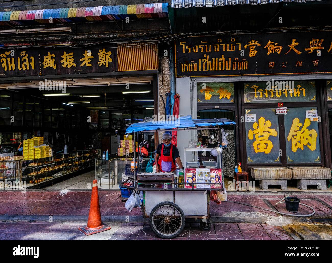 Ein männlicher Lebensmittelhändler steht hinter seinem Wagen vor einigen traditionellen chinesischen Unternehmen und Kulturgebäuden in Chinatown, Bangkok, Thailand. Stockfoto