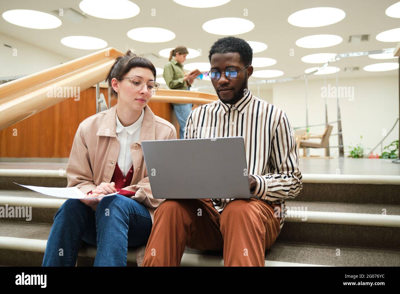 Afrikanischer Mann mit Laptop und studieren zusammen mit Frau, während sie auf Treppen in der Bibliothek sitzen Stockfoto