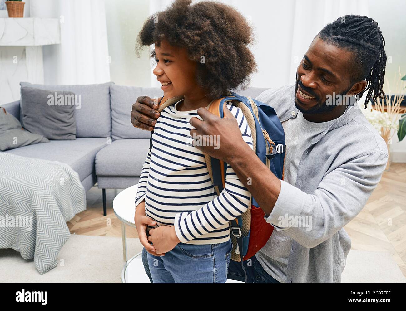 Glücklicher afroamerikanischer Vater hilft der Tochter, sich für die Schule vorzubereiten und legt ihr eine Schultasche auf den Rücken Stockfoto