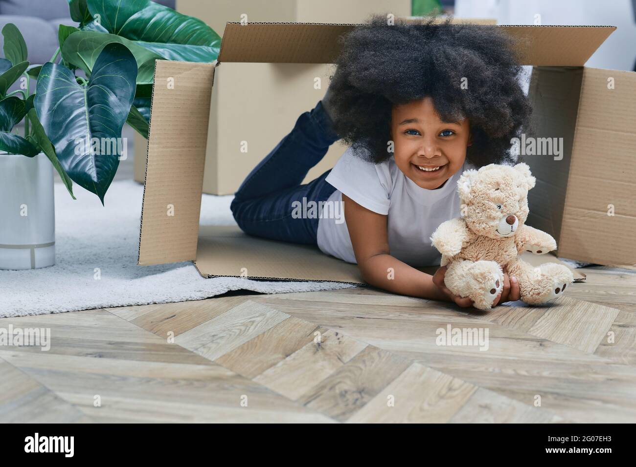 Glückliches afroamerikanisches kleines Mädchen, das mit Pappkartons und ihrem Plüschbären spielt und Spaß hat, während es nach Hause zieht Stockfoto