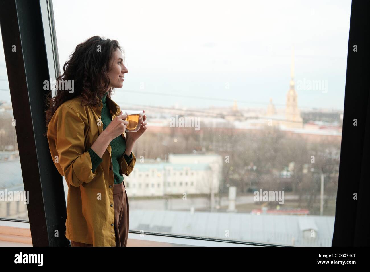 Junge Geschäftsfrau mit einer Tasse Tee und Blick durch das Fenster auf die Stadt, während sie im Büro steht Stockfoto