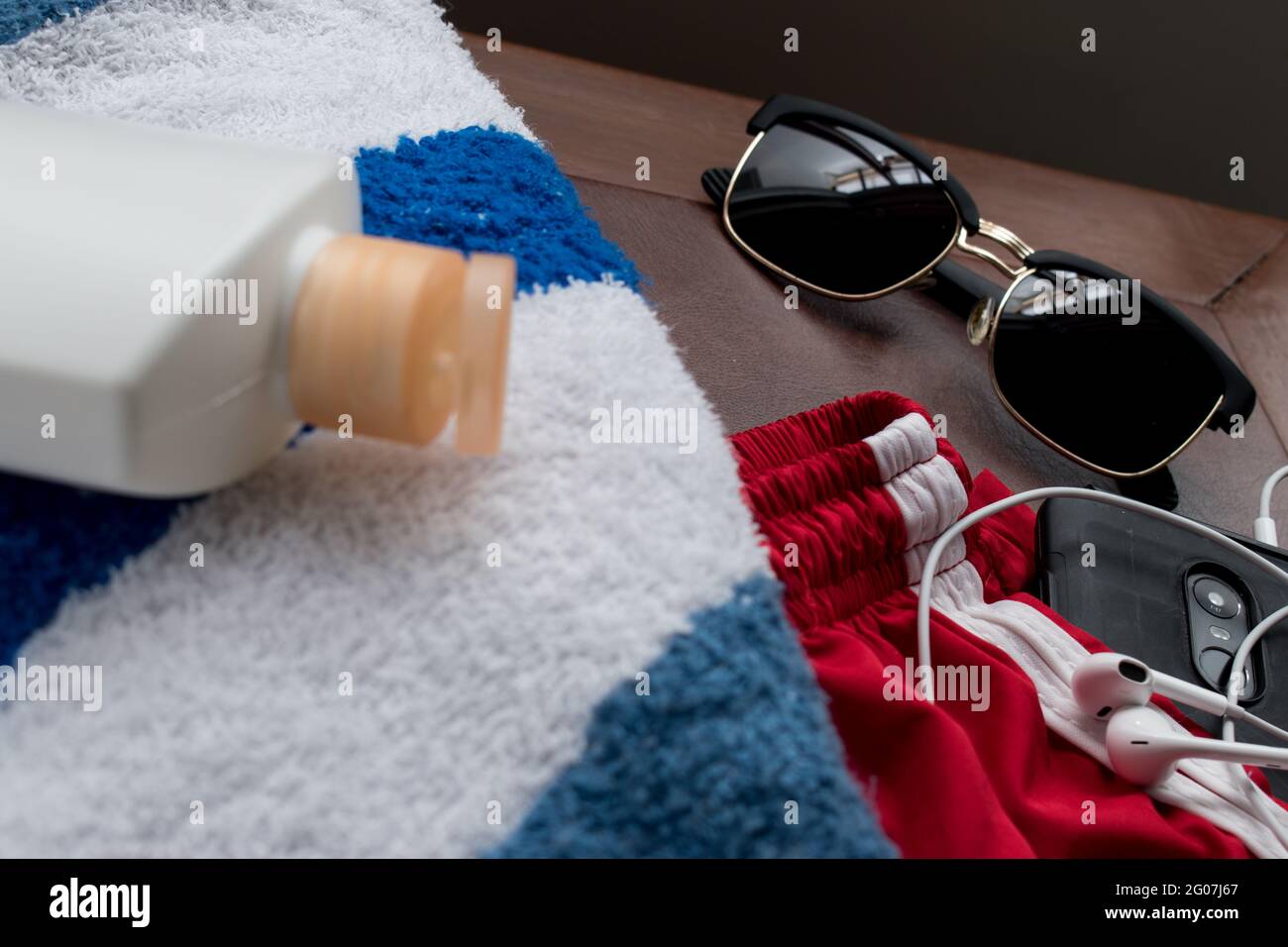 Blau-weiß gestreiftes Handtuch, cremefarbene Flasche, Sonnenbrille, schwarzes Smartphone, Kopfhörer und rote Hose für den Sommer auf einem braunen Holztisch Stockfoto