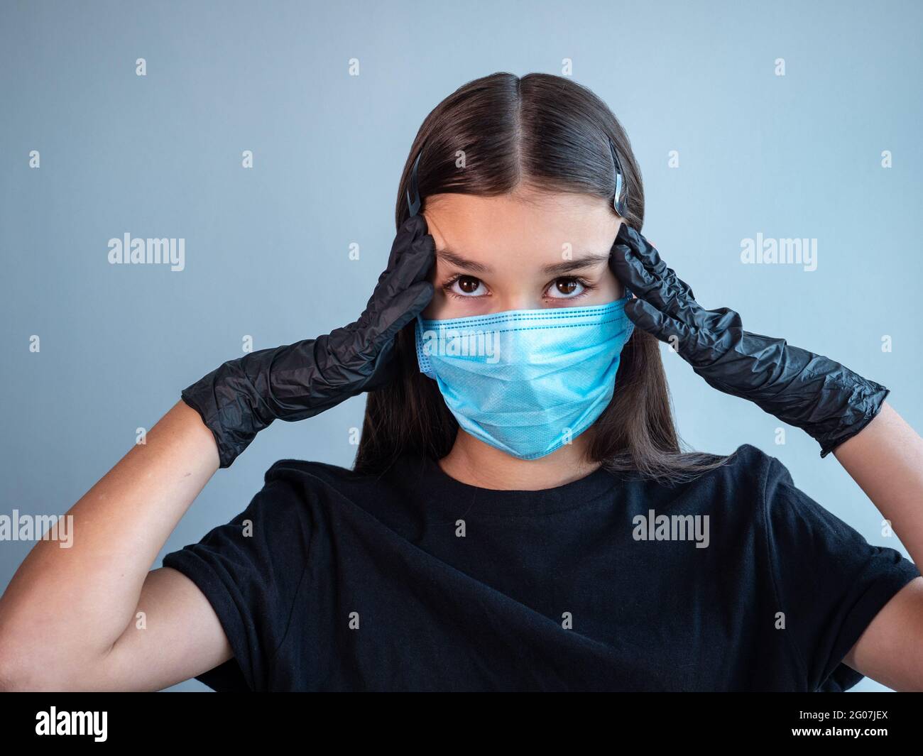 Ein Mädchen im Teenageralter in einer blauen medizinischen Schutzmaske berührt ihre Schläfen mit Händen in schwarzen Latexhandschuhen. Virales Infektionskonzept. Stockfoto