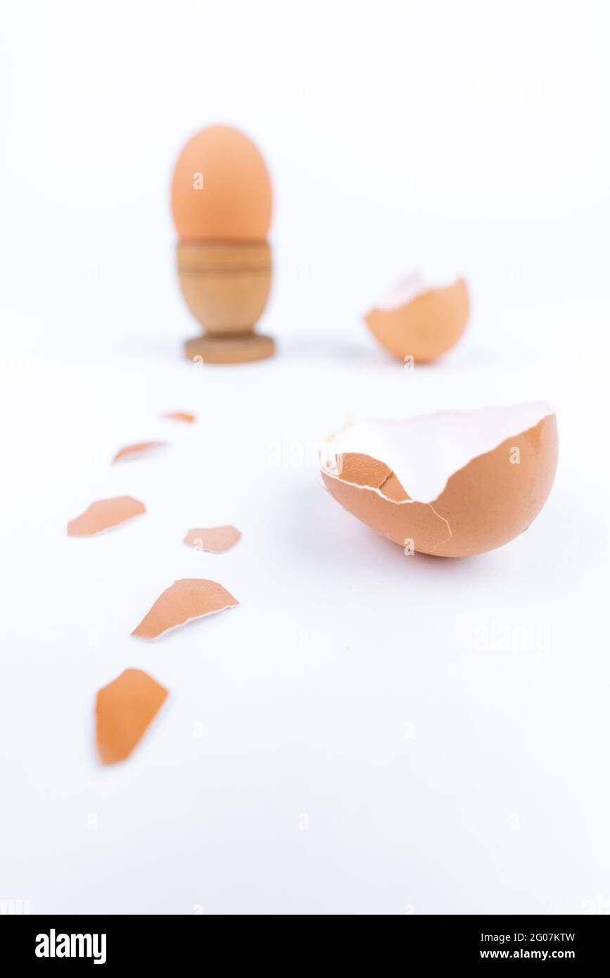 Vertikales Hühnereichen in altem hölzernen Eierstand und mehrere zerbrochene Eierschalen auf einer weißen Oberfläche. Minimalismus. Konzeptkunst für Lebensmittel oder tierische Produkte Stockfoto
