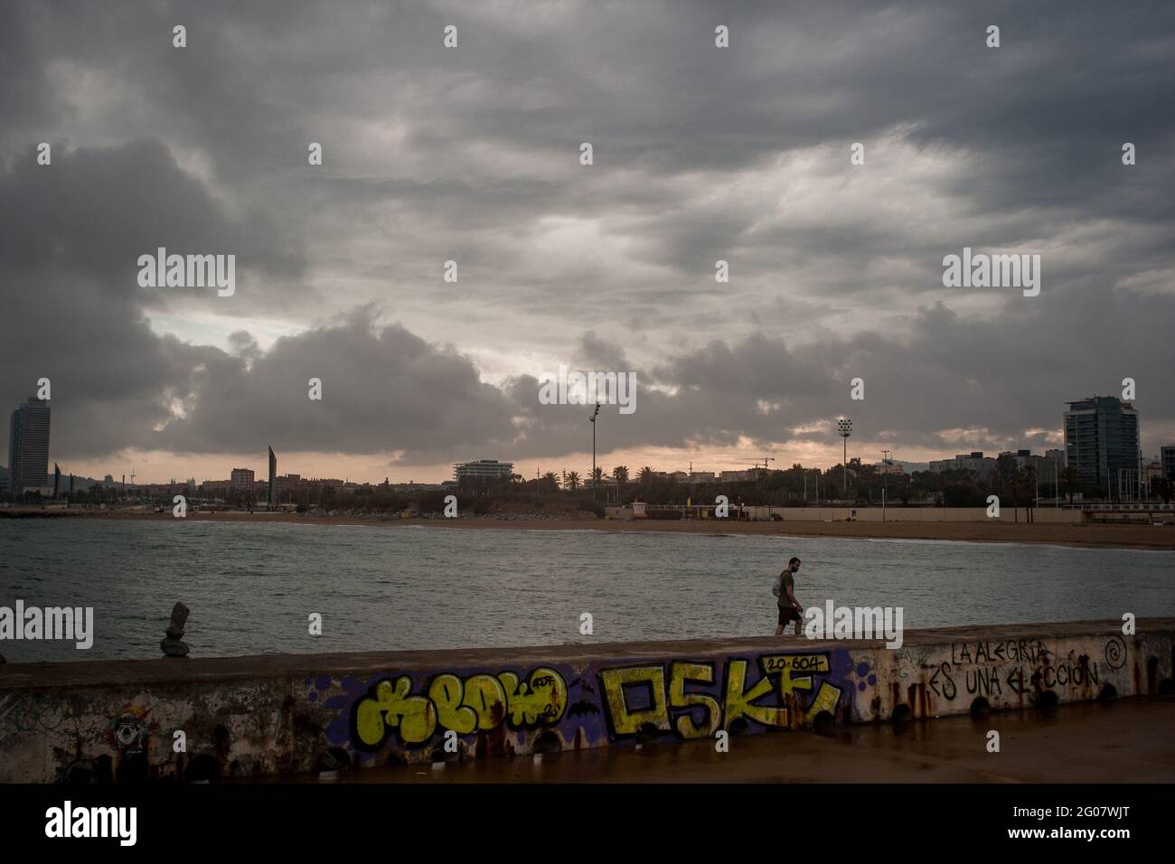 Ein Mann geht an einem Tag, der von regelmäßigen Regenschauern gekennzeichnet ist, an Barcelonas Küste entlang. Stockfoto