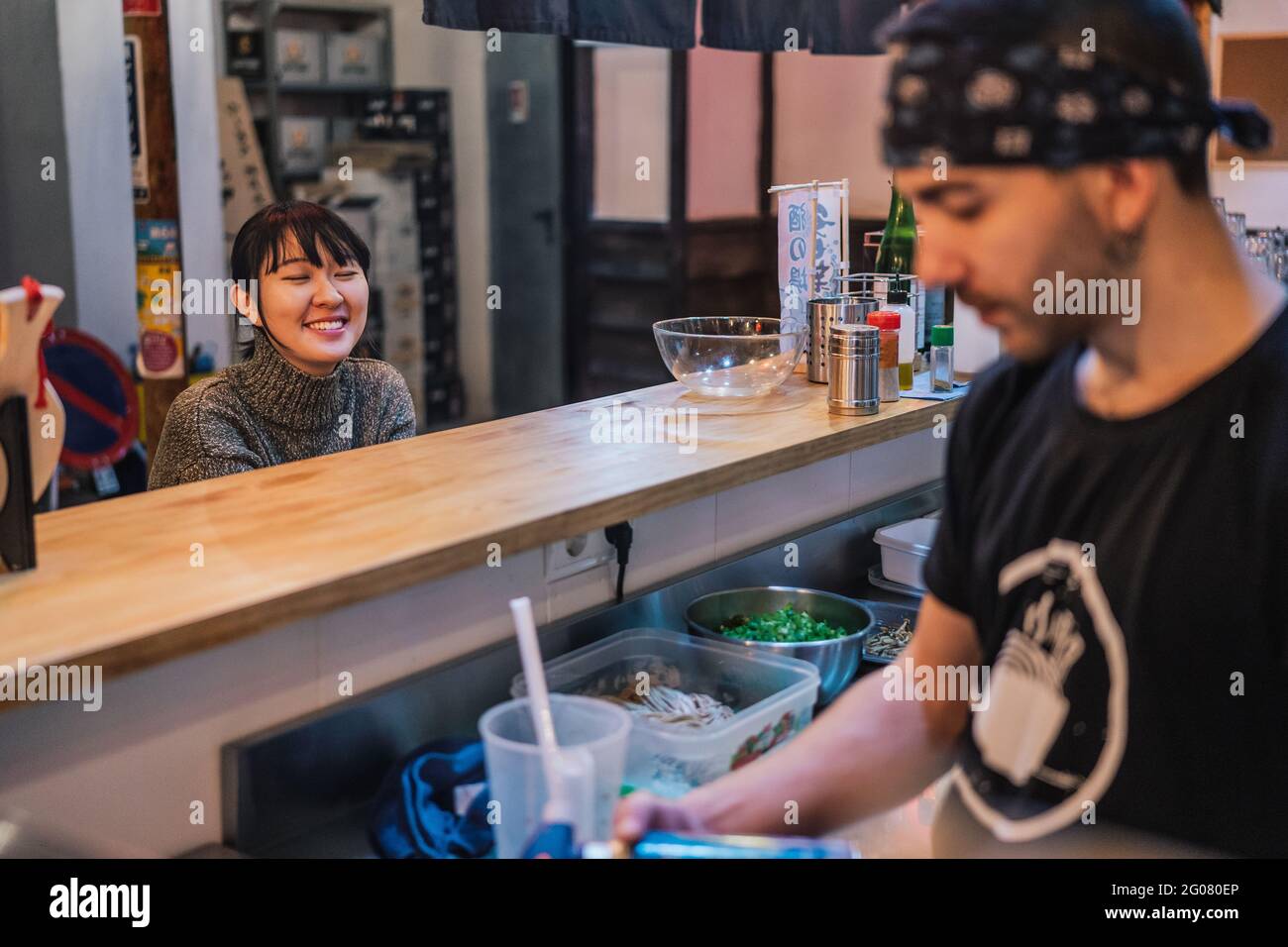 Asiatische Frau in legerer Kleidung sitzt an der Theke und spricht mit männlichen Arbeiter der modernen Ramen-Bar Stockfoto