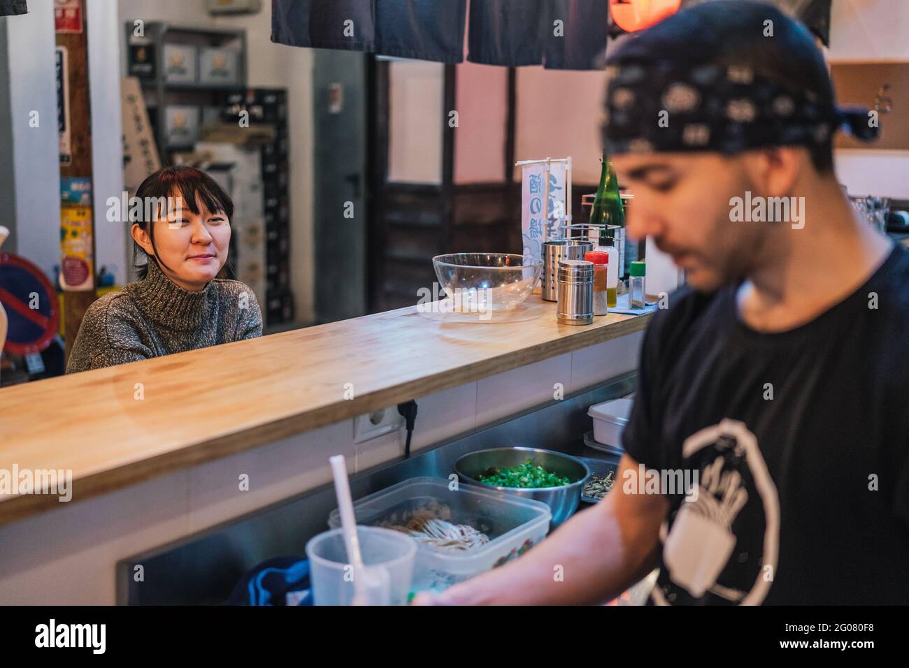 Asiatische Frau in legerer Kleidung sitzt an der Theke und spricht mit männlichen Arbeiter der modernen Ramen-Bar Stockfoto