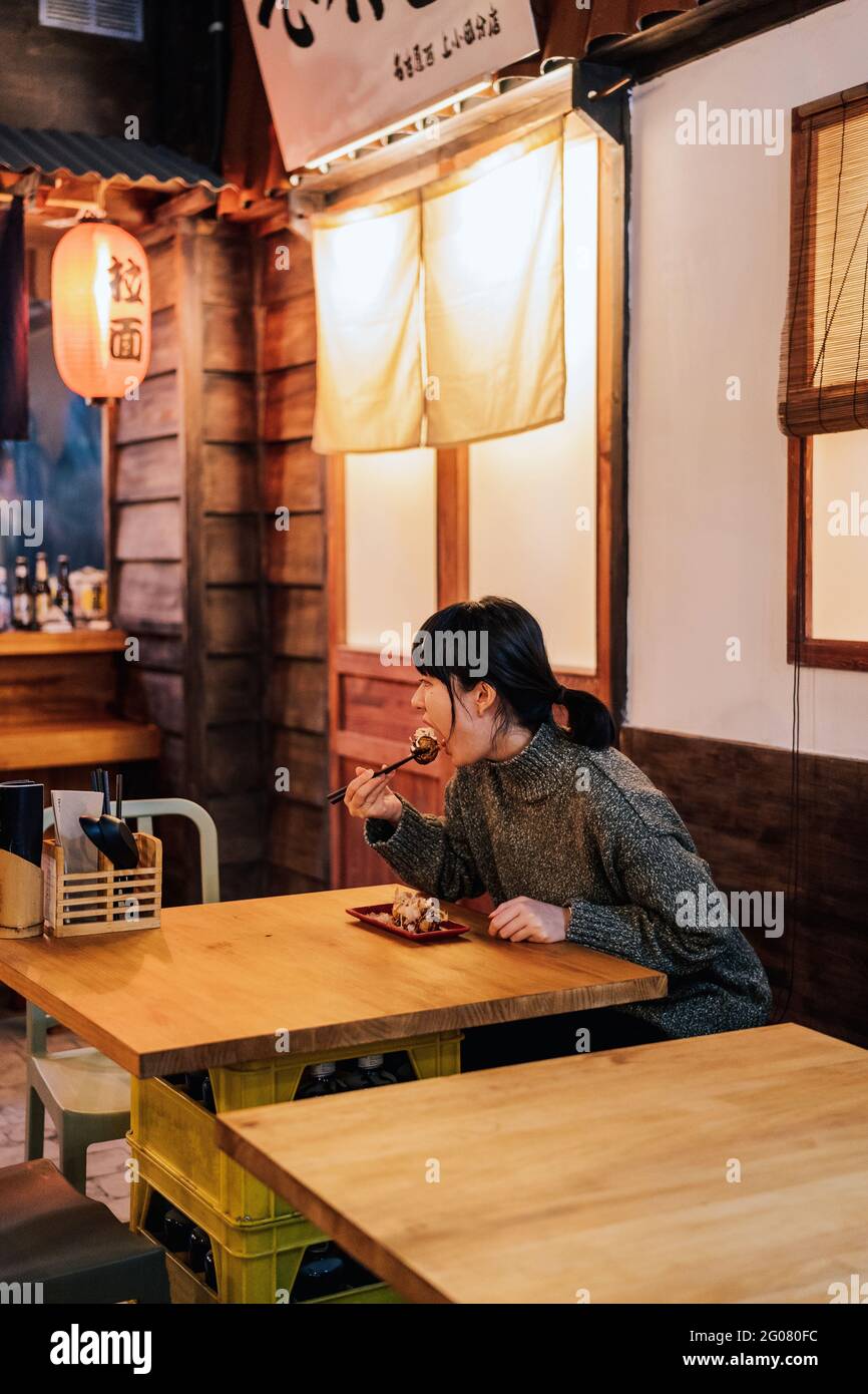 Junge asiatische Frau im Pullover, die an der Holztheke im Café isst Stockfoto
