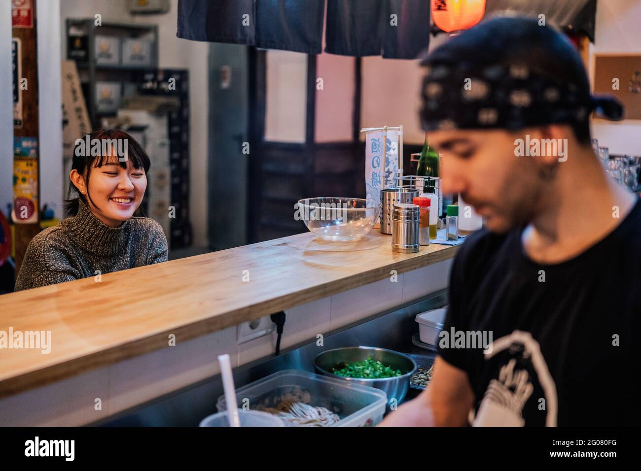 Asiatische Frau in legerer Kleidung sitzt an der Theke und spricht mit männlichen Arbeiter der modernen Ramen-Bar Stockfoto