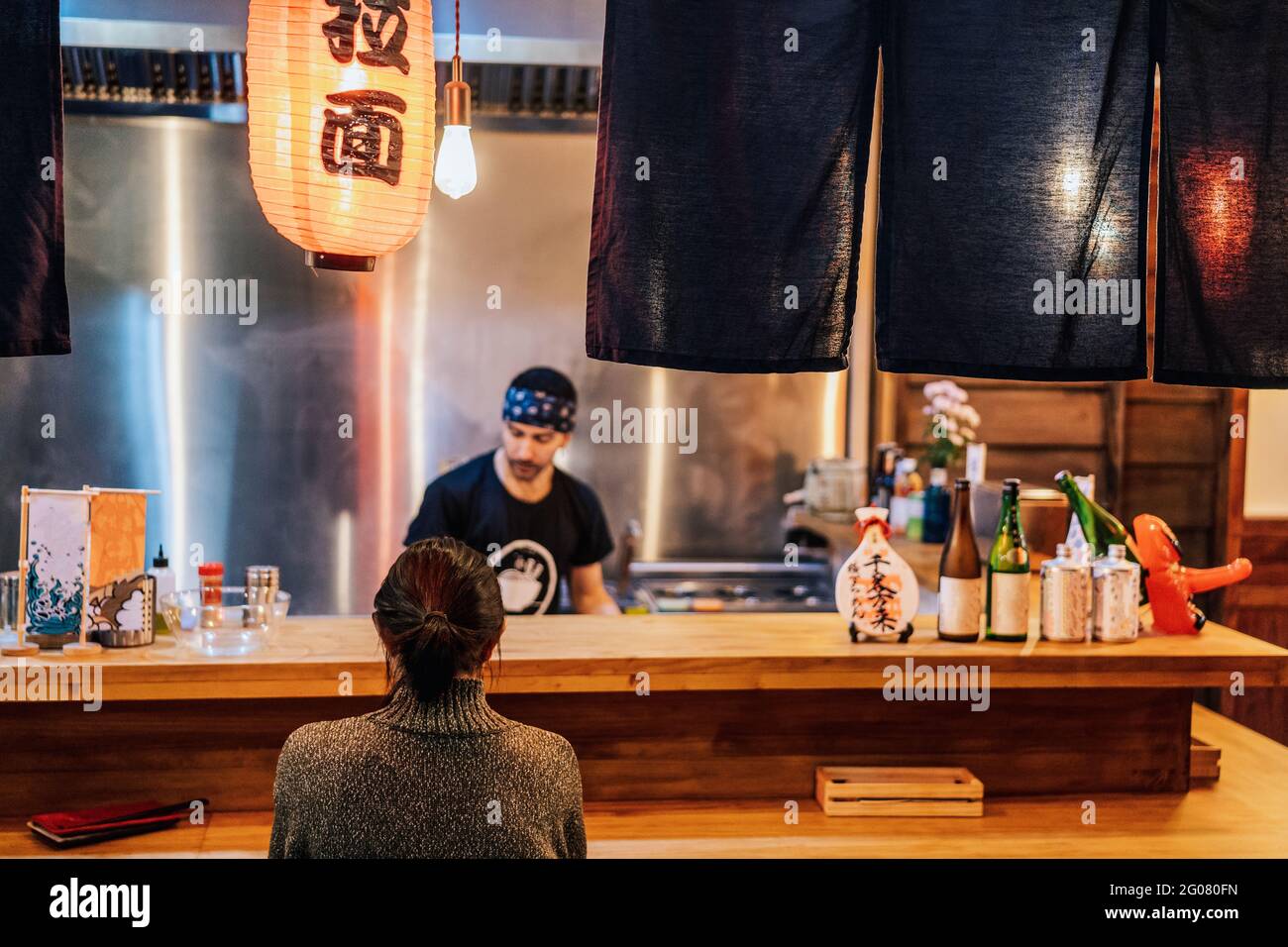 Rückansicht einer gesichtslosen, schwarzhaarigen Frau im Pullover, die an der Theke in der gemütlichen Ramen-Bar sitzt Stockfoto