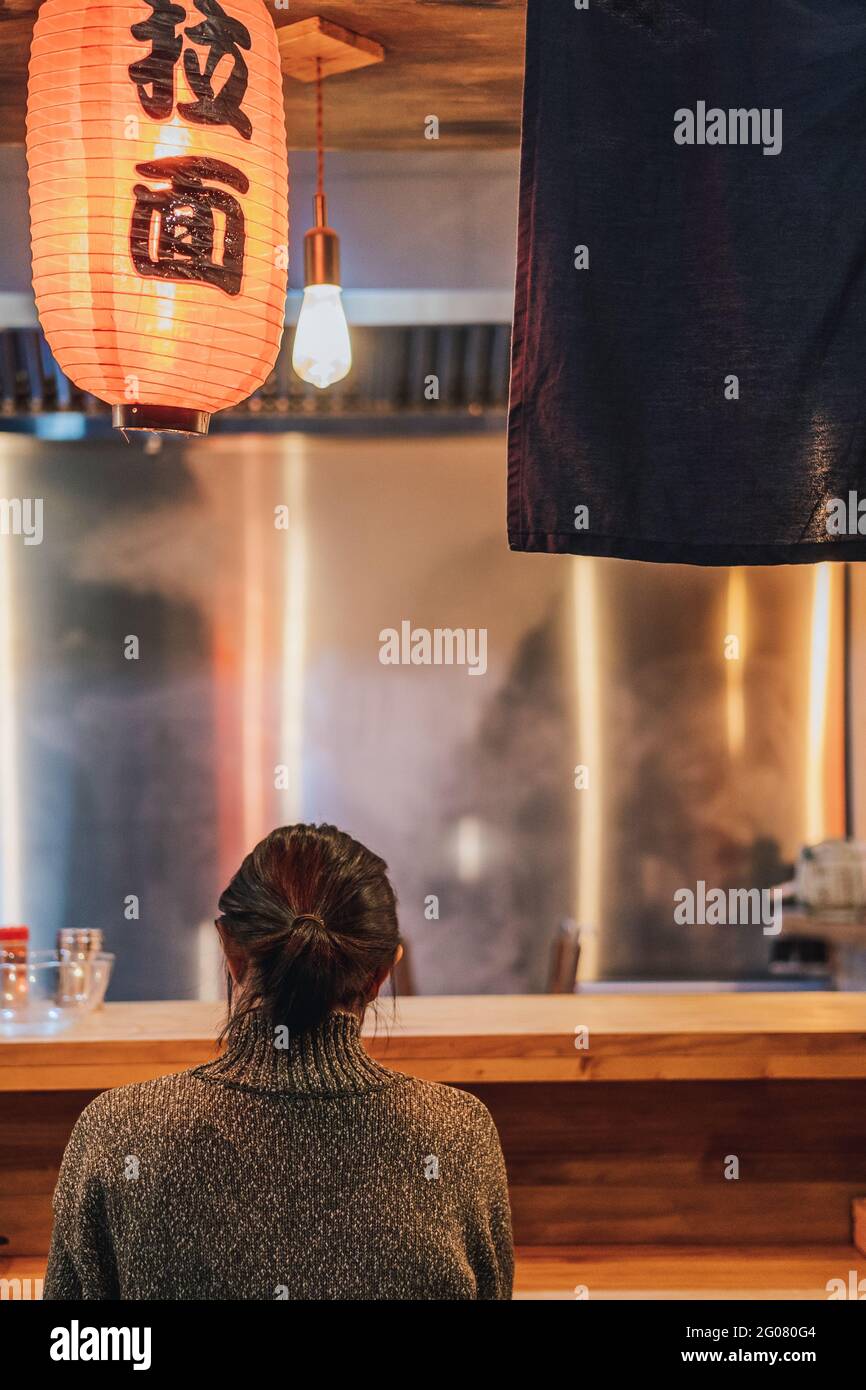 Rückansicht einer gesichtslosen, schwarzhaarigen Frau im Pullover, die an der Theke in der gemütlichen Ramen-Bar sitzt Stockfoto