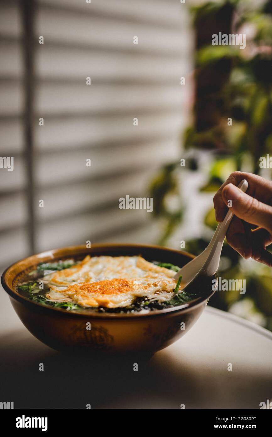 Hand einer anonymen Person, die Löffel verwendet, um leckere asiatische zu nehmen Suppe mit Nudeln und Spiegelei auf der Terrasse Stockfoto
