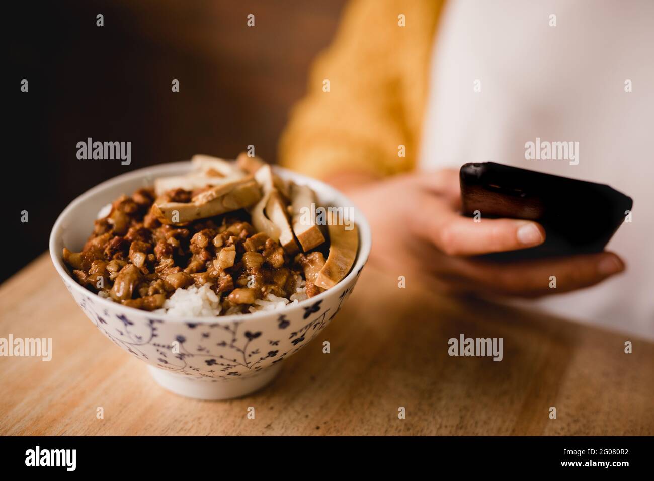 Schüssel mit traditioneller taiwanesischer Lu Rou Fächerschale aufgesetzt Holztisch in der Nähe weiblich mit Smartphone im Café Stockfoto