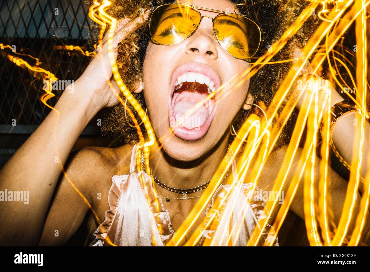 Ausdrucksstarke junge ethnische Dame mit Afro-Haaren in stilvoller Sonnenbrille Und oben berühren Kopf und laut schreiend, während sie sich abkühlen Nachtclub in der Nähe von Freeze l Stockfoto