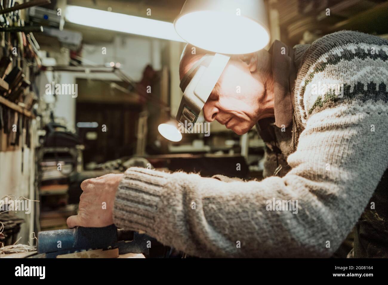 Seitenansicht des fokussierten älteren männlichen Geigenbauers in Pullover und Vergrößerungskopfgurt, der bei der Reparatur des Saiteninstruments an der Werkbank steht Detail in alter Stu Stockfoto