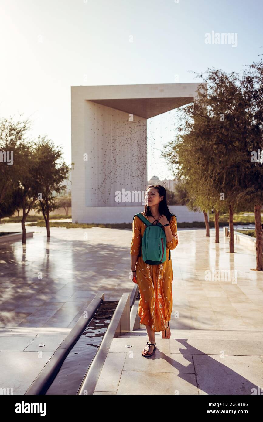 Ganzkörper positive junge weibliche Reisende in traditioneller Kleidung zu Fuß Auf einem gepflasterten Platz in der Nähe eines kubischen Pavillons am Gründerdenkmal In Abu Dhabi wil Stockfoto