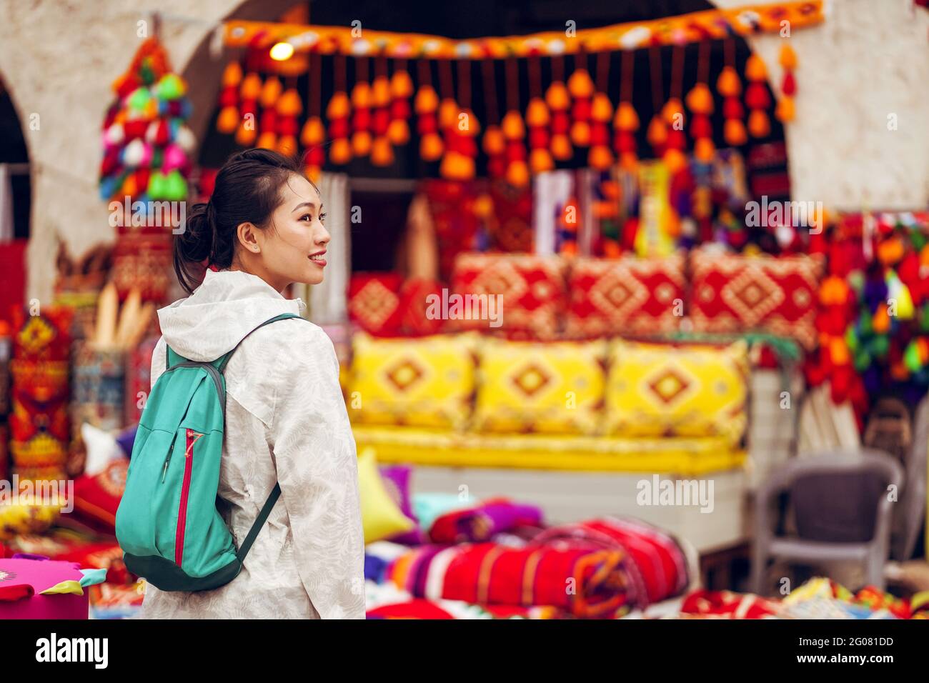 Rückansicht Zufriedene asiatische weibliche Touristin mit Rucksack lächelnd weg Während Sie sich gegen verschiedene traditionelle orientalische Kleidung und Souvenirs wehren Stockfoto