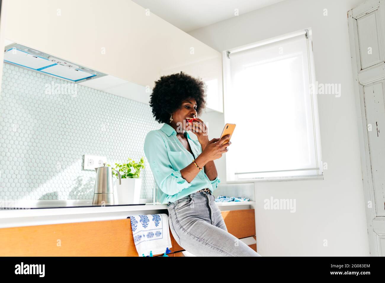 Von unten junge afroamerikanische Frau in lässiger Kleidung essen apple und Surfen im Internet auf dem Mobiltelefon, während Sie stehen Wohnküche Stockfoto