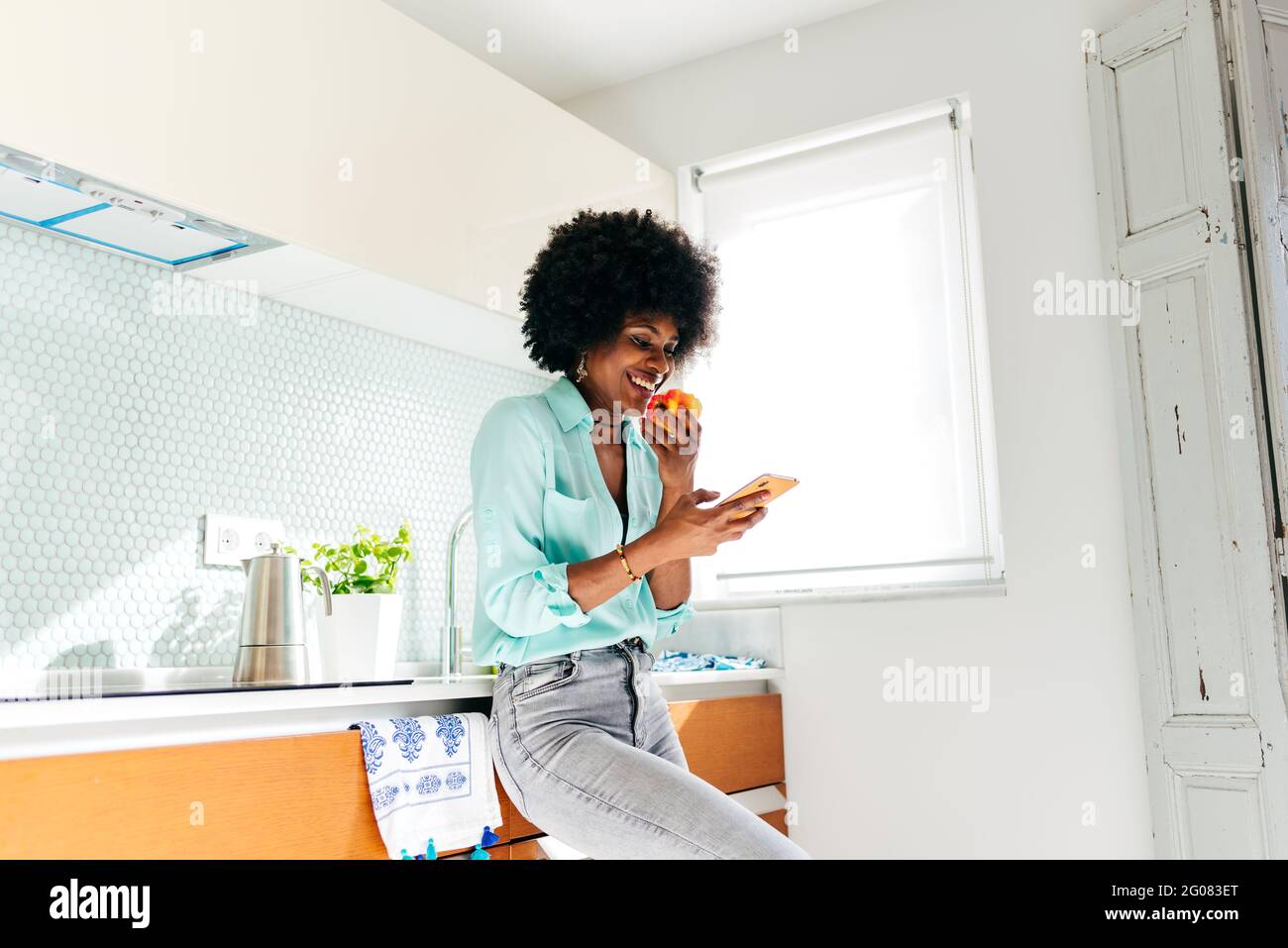 Von unten junge afroamerikanische Frau in lässiger Kleidung essen apple und Surfen im Internet auf dem Mobiltelefon, während Sie stehen Wohnküche Stockfoto