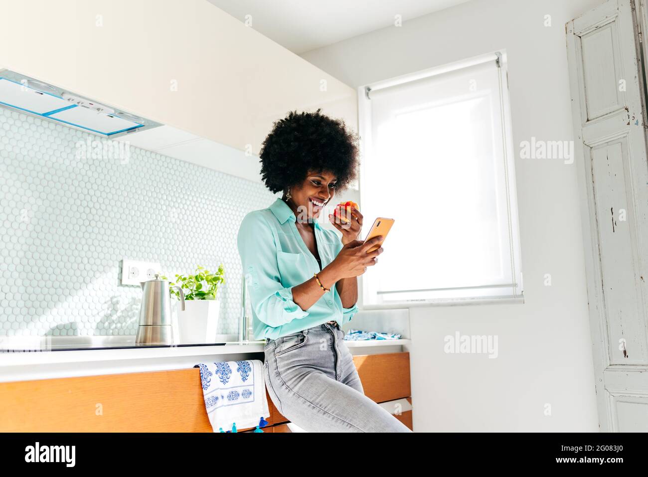 Von unten junge afroamerikanische Frau in lässiger Kleidung essen apple und Surfen im Internet auf dem Mobiltelefon, während Sie stehen Wohnküche Stockfoto