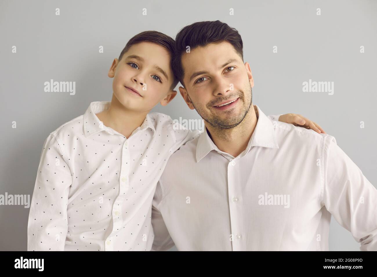 Vater mit Sohn, der in weißen Hemden gekleidet ist und kuschelt, während er auf grauem Hintergrund steht. Stockfoto