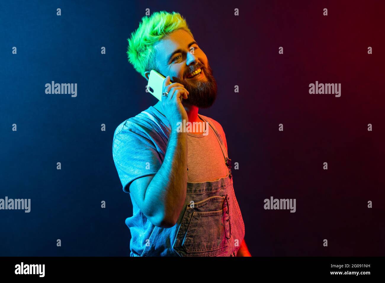 Porträt eines jungen erwachsenen Hipster-Mannes mit Denim-Overalls und T-Shirt-Sprechtelefon, hört glückliche Nachrichten, lächelt zahnig und drückt Optimismus aus. Farbenfroh Stockfoto