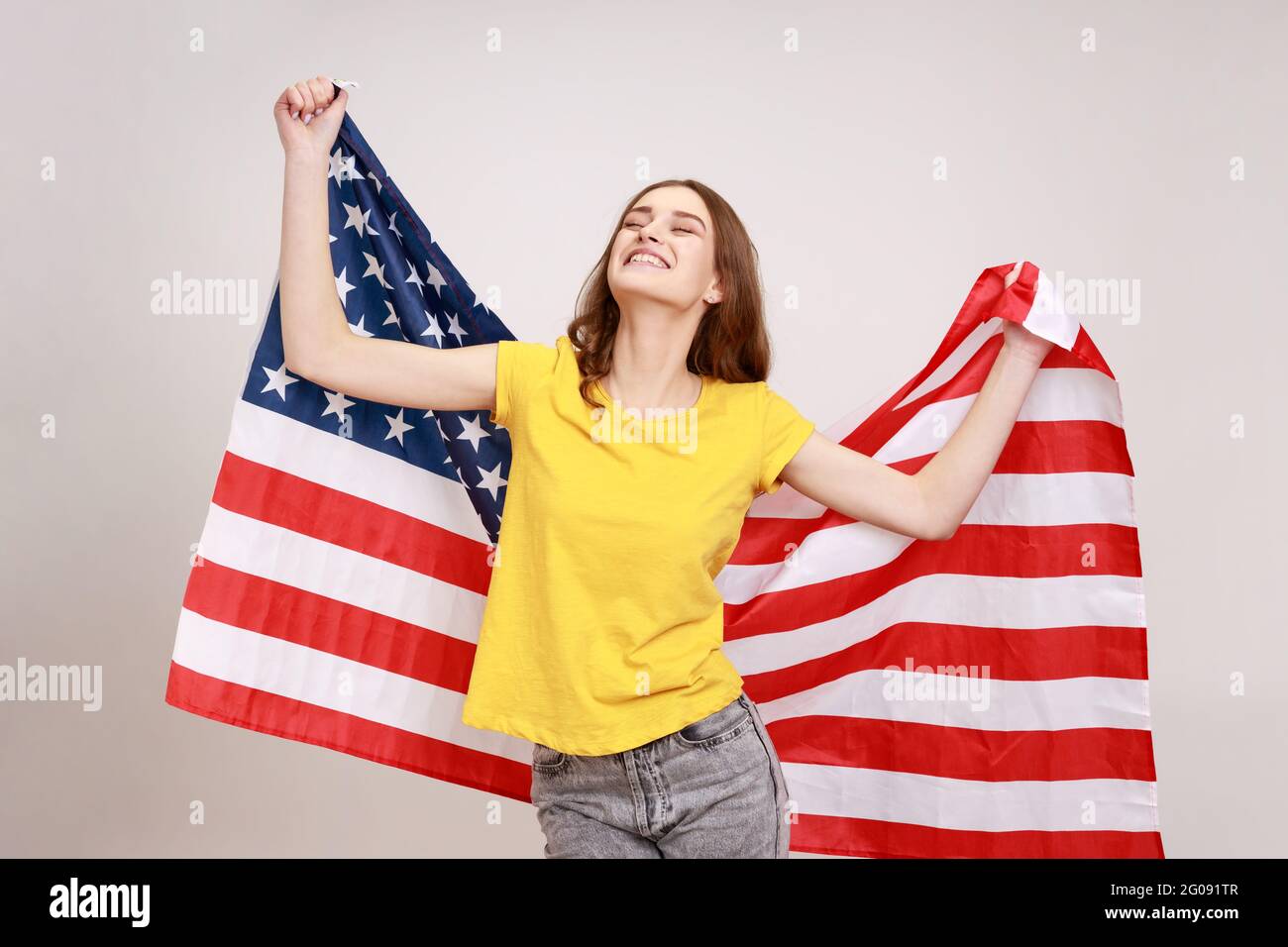Fröhliche, attraktive junge Frau mit welligen Haaren, gelbem T-Shirt mit der USA-Flagge über den Schultern, geschlossenen Augen und glücklichem Lächeln Stockfoto