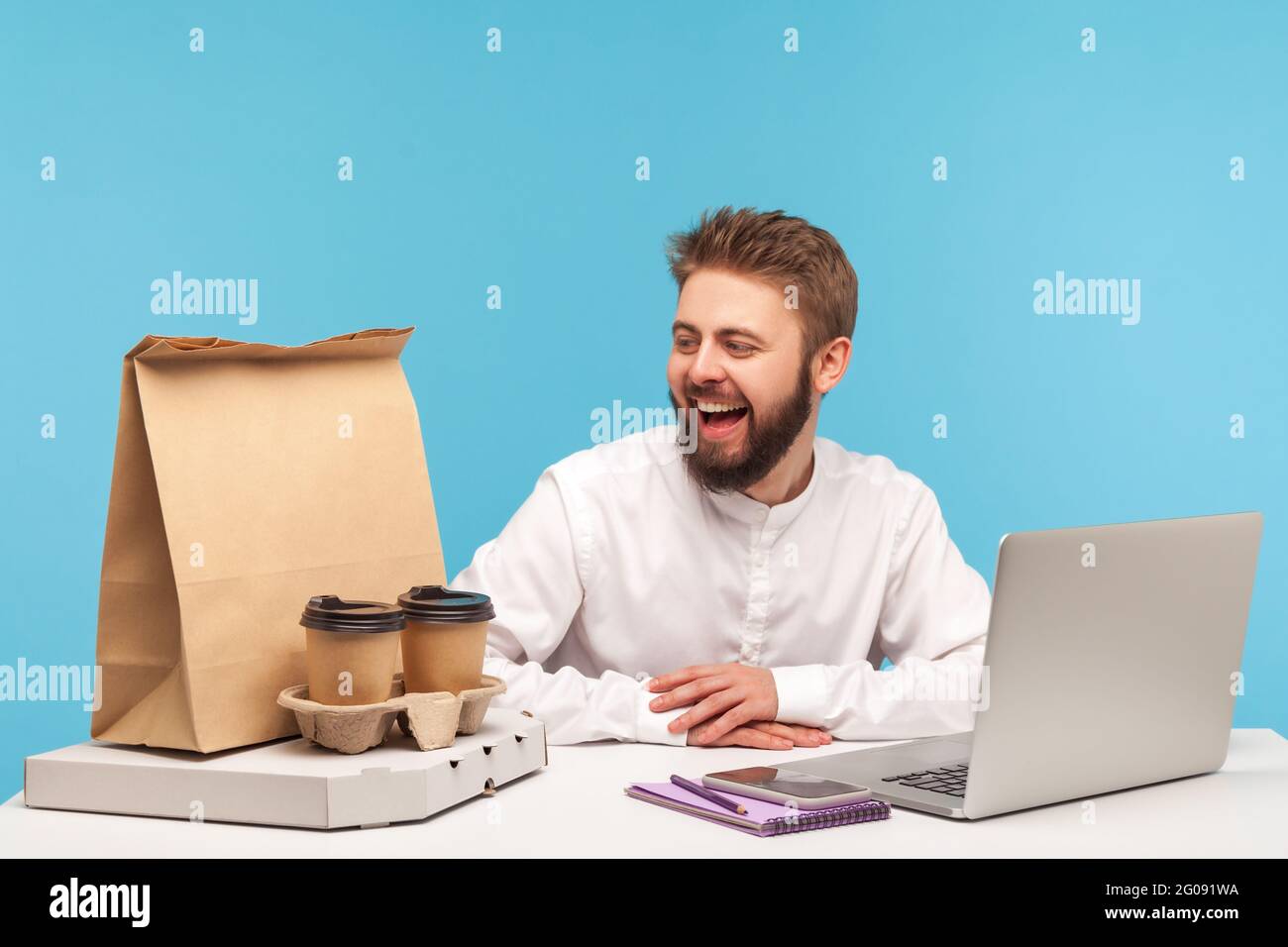 Glücklicher Büroangestellter, der mit einem Lächeln auf den Lippen die Pizzaschachtel, Tassen Kaffee und Papiertüte beim Kauf ansieht und die Fast Food-Lieferung genießt. Innenaufnahme des Studios i Stockfoto