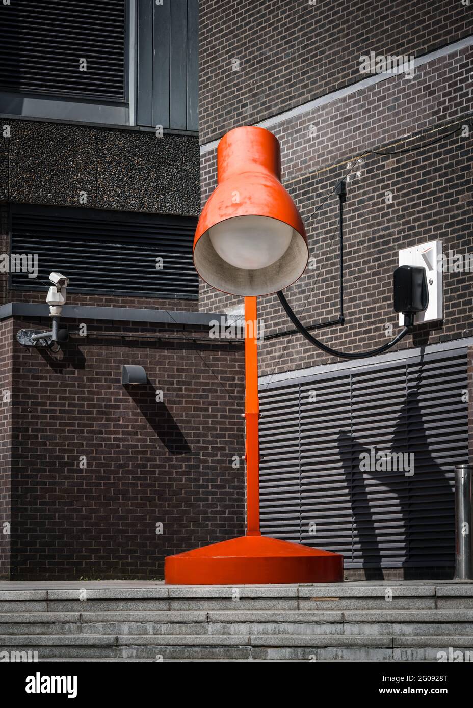 Riesige riesige, leuchtend rote Schreibtischlampe mit einer großen Lampe, die an der Außenwand des Gebäudes oben auf einer Treppe angeschlossen ist. Im Sommer hoch über der Ziegelmauer Stockfoto