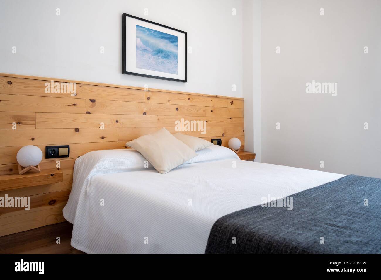 Die Einrichtung des hellen, weißen Schlafzimmers ist mit einem Foto der Meereswellen und einem Kopfteil aus Holz dekoriert Stockfoto