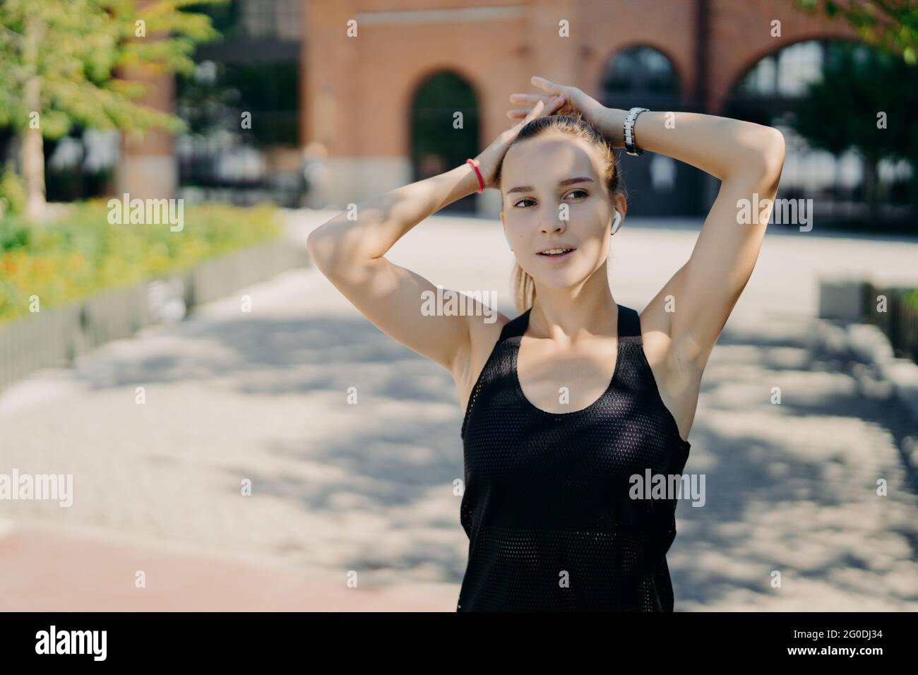 Die sportliche Frau, die sich in die Ferne konzentriert, atmet nach dem Joggen während des Cardio-Trainings tief ein, gekleidet in schwarzes T-Shirt hält die Hände am Kopf Stockfoto