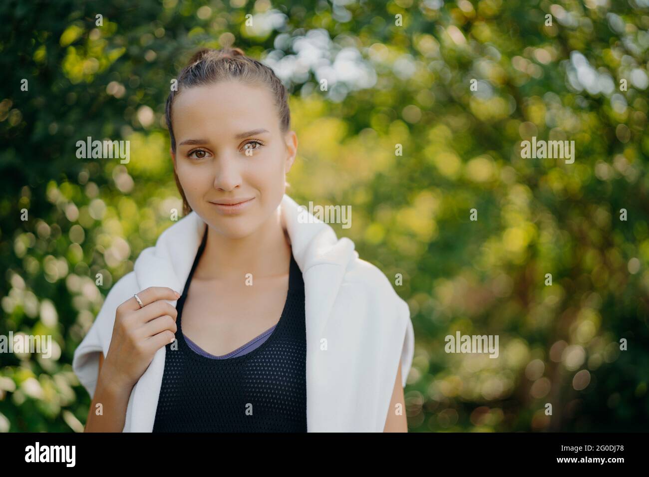 Schöne dunkelhaarige Europäerin trägt schwarzes T-Shirt und weißen Pullover über den Schultern schaut direkt auf die Kamera hat regelmäßige Outdoor-Training Spaziergänge in Stockfoto