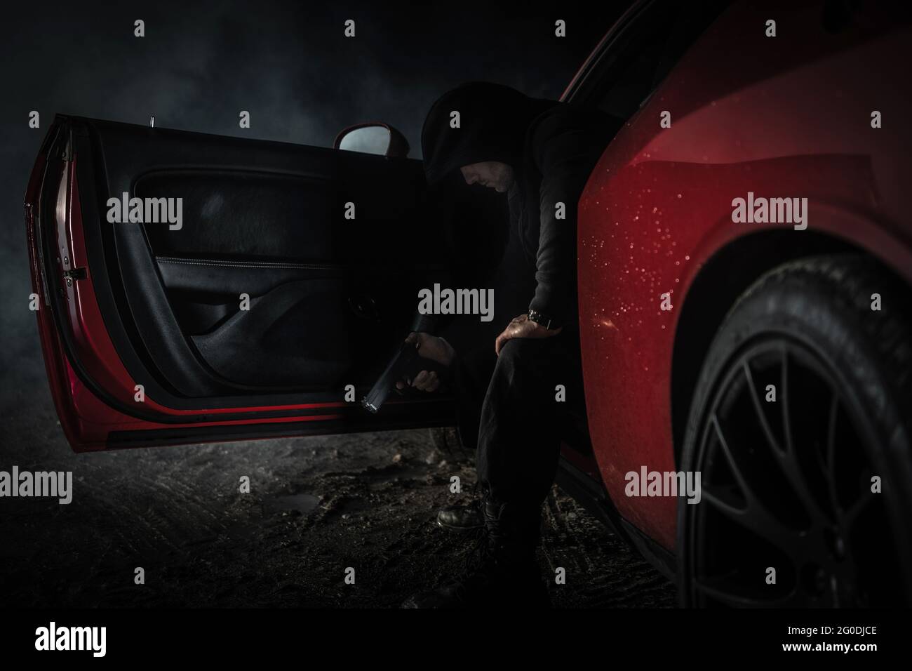Nachdenklicher kaukasischer Gangster in den 40er Jahren mit schwarzem Hoodie und Handfeuerwaffe in der Hand im Red Muscle Car. Unzulässiges Aktivitätsprinzip. Stockfoto
